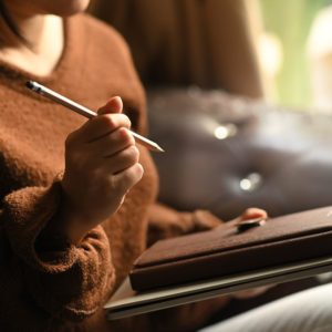 Woman holding a pencil and notebook in her hands while taking notes and sitting at the leather couch over comfortable living room as background.